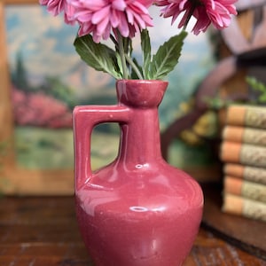 May include: A pink ceramic vase with a handle, holding a bouquet of pink flowers.
