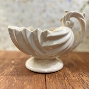 May include: Cream-colored, vintage ceramic bowl with a pedestal base and a decorative, swirling handle. The bowl has a scalloped rim and a textured, ribbed design. The piece is displayed on a wooden surface.