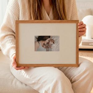 Può includere: Una cornice rettangolare in legno con un passe-partout chiaro. La cornice contiene una fotografia di una madre e un bambino. La cornice è tenuta da una persona che indossa un maglione e pantaloni color crema.