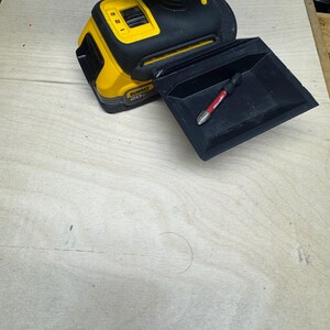 May include: A yellow and black cordless drill with a black accessory tray. A red and silver drill bit is in the tray. The drill is on a light wood surface.