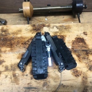 以下が含まれることがあります： 黒いマルチツールで、銀色の刃と黒いハンドルが付いています。ツールは開いており、レンチ、ドライバー、ペンチなど、さまざまなアタッチメントが付いています。ツールは木製の表面の上に置かれています。