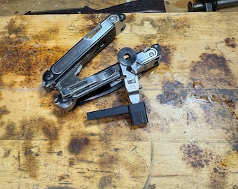 May include: A silver multi-tool with a black handle and a variety of tools, including a knife, pliers, and a screwdriver. The multi-tool is open and the tools are visible. The multi-tool is lying on a wooden surface.