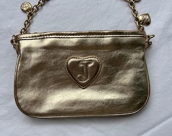 May include: A gold-colored clutch with a heart-shaped emblem featuring the letter "J". The clutch has a gold chain handle adorned with heart and charm accents. The bag is made of a shiny, metallic material.