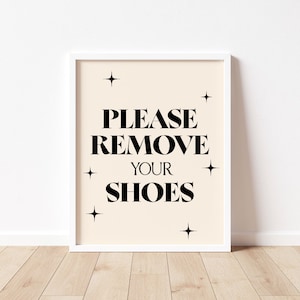 May include: White framed print with a beige background and black text that reads "Please Remove Your Shoes" with small black stars around the text.