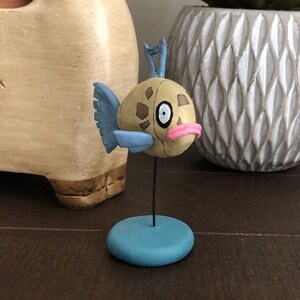 Puede incluir: Una figurita pintada a mano de un pez marrón y azul con una boca rosa. El pez está de pie sobre una base azul y tiene un soporte de metal negro.