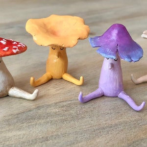 Może przedstawiać: Cztery ręcznie robione ceramiczne figurki grzybów z różnokolorowymi kapeluszami, siedzące w rzędzie ze skrzyżowanymi nogami. Grzyby są pomalowane w odcieniach czerwonego, żółtego, fioletowego i brązowego.