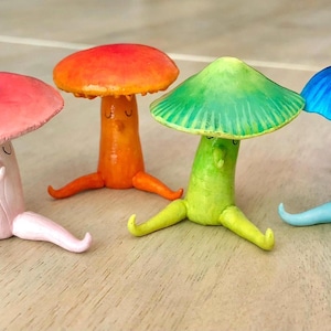 May include: Four whimsical mushroom figurines in pink, orange, green, and blue. Each has a rounded cap, a stem with a smiling face, and legs. The colorful mushrooms are arranged on a light brown surface.