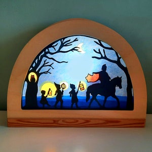 May include: A wooden silhouette light-up scene with a blue background, depicting a knight on horseback leading a group of children carrying lanterns through a forest. The scene is illuminated from behind, creating a warm glow.