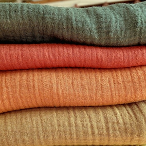 Puede incluir: Una pila de cuatro piezas dobladas de tela de muselina texturizada. La capa superior es de color verde bosque profundo, seguida de un rojo coral vibrante, un naranja calabaza cálido y una capa inferior de color amarillo mostaza apagado.