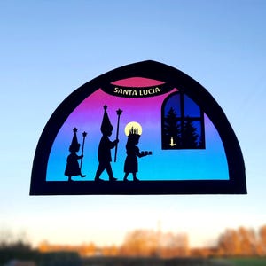 May include: A colourful stained glass window decoration featuring a procession of three figures in silhouette, carrying candles and stars. The window is shaped like a half-circle and has a blue and purple gradient. The text "SANTA LUCIA" is written in yellow at the top of the window.