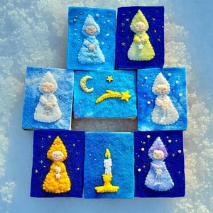 Könnte beinhalten: Neun Filzkunstwerke auf einer schneebedeckten Oberfläche. Jedes Stück zeigt ein anderes Design, darunter Figuren in Hüten und Gewändern, einen Halbmond, einen Sternschnuppen und eine Kerze. Die Stücke sind hauptsächlich blau, gelb und weiß.