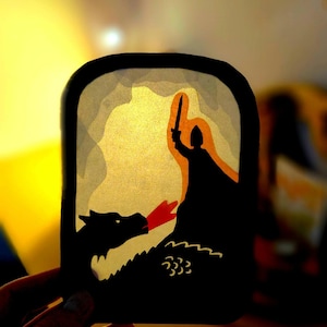 May include: A backlit paper cutout art piece featuring a black silhouette of a knight holding a sword aloft, facing a dragon breathing red fire. The scene is framed in a rounded rectangular border with layers of yellow and white light behind it.