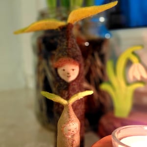 May include: A whimsical, handmade felt figure with a smiling face and yellow leaf-like accents. The figure is light brown with a stitched design. A larger figure with a similar design is in the background, along with a candle and a glass jar.