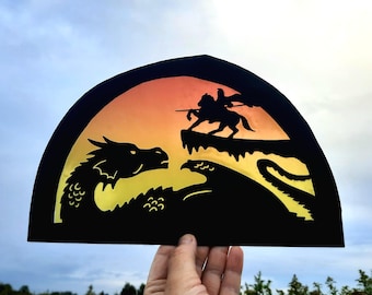 May include: A black paper silhouette of a dragon and a knight on horseback against a sunset background. The silhouette is cut out of a black paper circle.