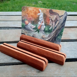 May include: Three wooden picture stands with a brown finish. The stands are stacked on top of each other. A card with a watercolour illustration of a rabbit, a squirrel, and a hedgehog is displayed in the stands.