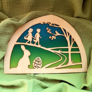 Può includere: Scena pasquale in legno con due bambini che camminano lungo un sentiero, un coniglio seduto vicino a un uovo decorato e un albero con un uccello su un ramo. Lo sfondo è un gradiente di blu, verde e giallo.