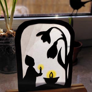 May include: A black silhouette of a child holding a candle, with a candlelit bowl and floral elements. The artwork is set in a black frame on a wooden base. A bird silhouette is visible in the background.