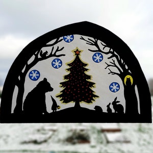 Puede incluir: Silueta negra de un árbol de Navidad con adornos rojos y amarillos, un oso, un conejo y un búho en un paisaje invernal nevado. El árbol está rodeado de copos de nieve azules.
