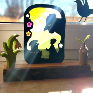 Könnte beinhalten: Ein Holzregal zeigt eine schwarze Silhouettenszene eines Vogels, einer Sonne und Figuren mit bunten Blumenakzenten. Eine grüne Holzpflanze und eine Filzpuppe mit einem Pflanzenspross befinden sich auf beiden Seiten. Die Szene spielt sich vor einem Fenster ab.