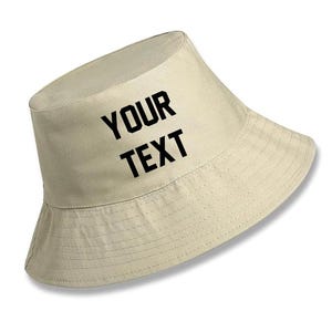 May include: A beige bucket hat featuring the black text YOUR TEXT printed on the front crown. The hat has a flat top, a structured crown, and a downward-sloping brim with visible stitching details around the circumference of the brim.