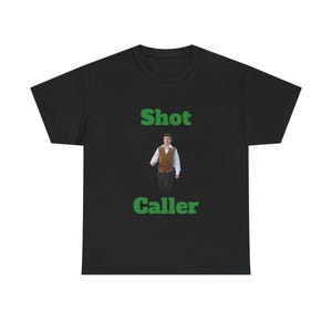 May include: Black t-shirt with the words "Shot Caller" in green block letters. A small image of a person wearing a vest and dress shirt is centered between the words. The shirt is made of a soft material.