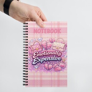 May include: A pink spiral-bound notebook with the word "NOTEBOOK" at the top. The cover features illustrations of handbags, broken hearts, diamonds, and perfume bottles. The phrase "Emotionally Expensive" is written in cursive.