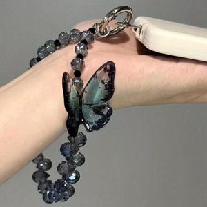 May include: A beaded keychain with a butterfly charm. The keychain features a silver-colored clasp and a strand of faceted, gray beads. The butterfly charm has a gradient of dark blue and green hues. A white rectangular object is attached to the clasp.
