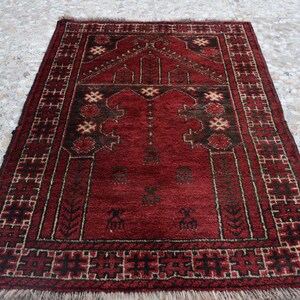 May include: A red prayer rug with a traditional geometric pattern and a central design. The rug has a fringe border and is made of wool.