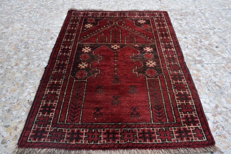 May include: A red prayer rug with a traditional geometric pattern and a central design. The rug has a fringe border and is made of wool.