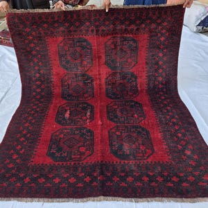 May include: A rectangular, red rug with a central design of eight black geometric shapes. The rug has a black border with repeating floral patterns and a fringed edge. The rug is held up by several people.