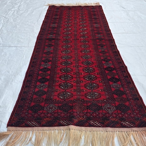 May include: A rectangular, red rug with a detailed, geometric pattern in black. The rug has a central design of repeating oval shapes and diamond patterns. The edges are finished with a fringe.