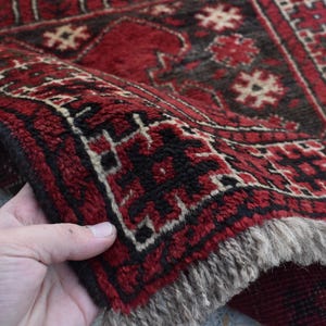 May include: A red and black patterned rug with a fringed border. The rug is folded in half and held by a hand.