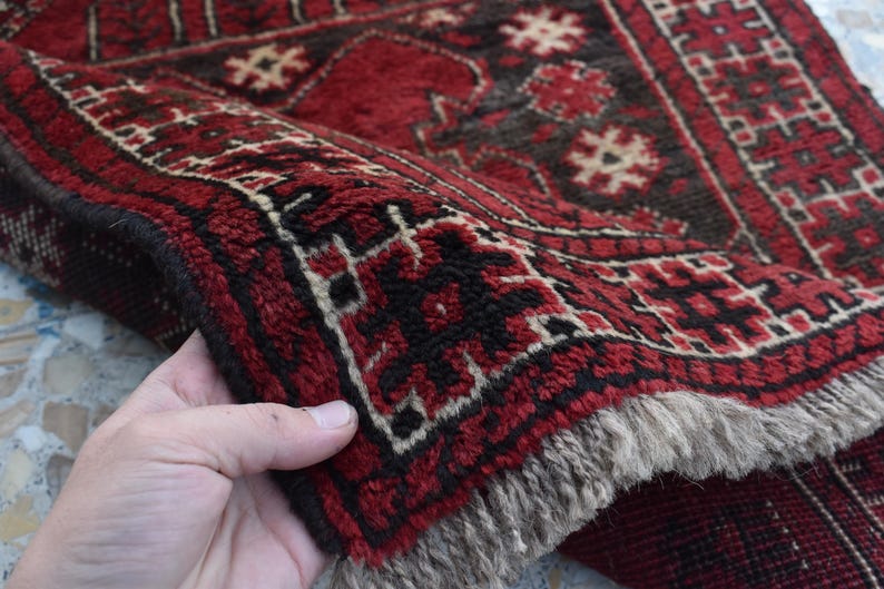 May include: A red and black patterned rug with a fringed border. The rug is folded in half and held by a hand.