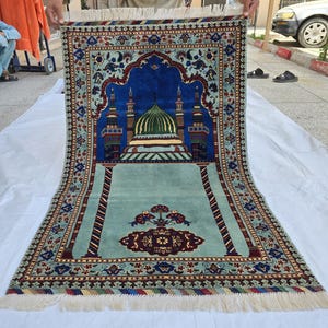 May include: An intricately designed prayer rug featuring a mosque with a green dome and blue accents. The rug has a light green field with a detailed border in shades of red, blue, and gold. The rug is rectangular with a fringed edge.