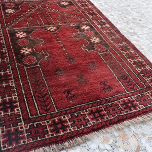 May include: A red prayer rug with a traditional geometric pattern and a central design featuring a mosque arch. The rug has a fringe border and is made of wool.