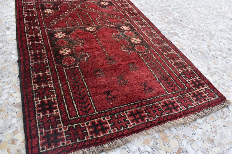 May include: A red prayer rug with a traditional geometric pattern and a central design featuring a mosque arch. The rug has a fringe border and is made of wool.