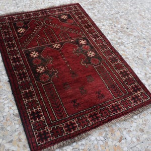 May include: A red prayer rug with a traditional geometric pattern and intricate details. The rug has a fringed edge and is made of wool.