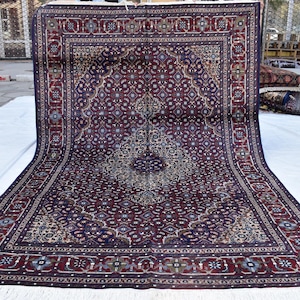 May include: A traditional Persian rug with a dark blue background and intricate floral patterns in shades of red, cream, and beige. The rug has a border with a repeating geometric design.