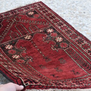 May include: A red prayer rug with a traditional geometric pattern and a central design. The rug has a fringe border and is made of wool.