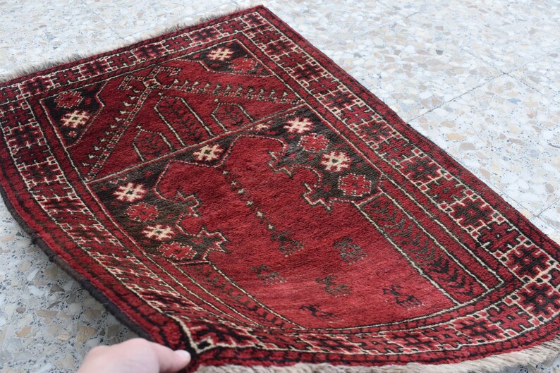 May include: A red prayer rug with a traditional geometric pattern and a central design. The rug has a fringe border and is made of wool.
