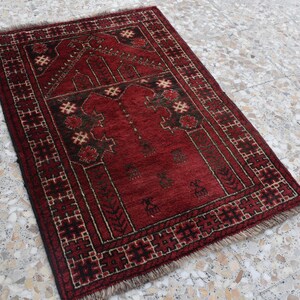 May include: A red prayer rug with a traditional geometric pattern and intricate details. The rug has a fringed edge and is made of wool.