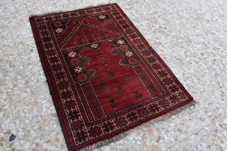 May include: A red prayer rug with a traditional geometric pattern and intricate details. The rug has a fringed edge and is made of wool.