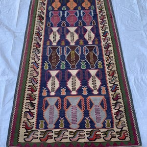 May include: A vintage Turkish rug with a geometric pattern featuring stylized vases in shades of blue, pink, orange, and green. The rug has a cream-colored background and a fringe border.