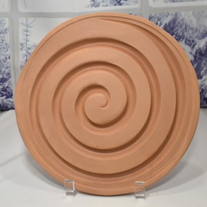 May include: A terracotta-colored round plate with a raised spiral design. The plate is made of clay and has a rustic look.