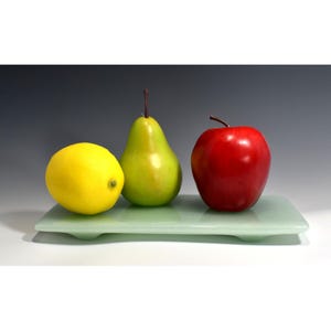 May include: A green pear, a yellow lemon, and a red apple are arranged on a light green glass serving tray.