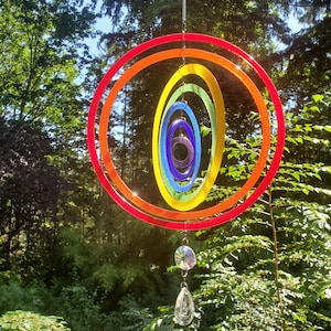 May include: A colorful, rainbow-themed wind spinner with concentric, oval-shaped rings in red, orange, yellow, green, blue, and purple. The wind spinner is suspended by a chain and features a crystal pendant. The background is a lush green forest.