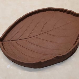 May include: A brown clay leaf-shaped dish with a textured surface. The dish is designed to resemble a leaf with veins and a scalloped edge.