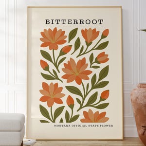 Puede incluir: Una impresión artística enmarcada con una ilustración botánica de la flor Bitterroot, la flor oficial del estado de Montana. La obra de arte muestra flores y capullos de color naranja con hojas verdes sobre un fondo crema. La palabra "BITTERROOT" está en la parte superior.