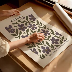 A person's hands are holding a poster with a floral design featuring purple flowers and green leaves.