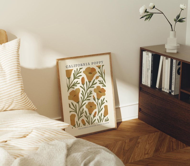 a framed poster of California poppies on a bedside table, with a bookshelf and a vase of white flowers in the background.